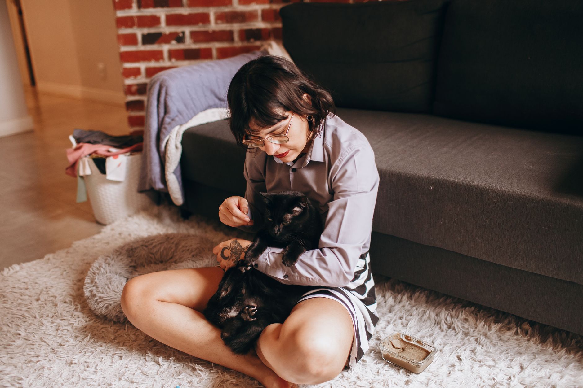 A woman lovingly pets her two black cats while sitting on a soft rug in a cozy living room.